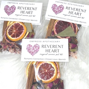 May include: Three clear pouches containing dried orange slices, cinnamon sticks, rose petals, and other botanicals. Each pouch has a label that reads "REVERENT HEART magical simmer pot kit" with the words "Romantic Connection, Chosen Love, Warmth."