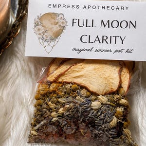 May include: A clear plastic pouch containing dried herbs, flowers, and fruit slices with a label reading Empress Apothecary Full Moon Clarity magical simmer pot kit, next to a lit candle in a glass jar and a long, translucent crystal wand.