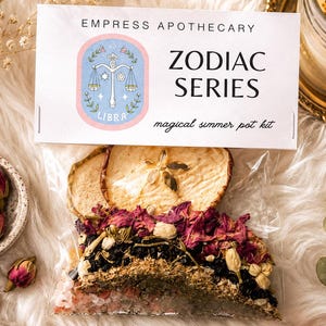May include: A clear plastic pouch containing a simmer pot kit with dried apple slices, pink rose buds, dark berries, and herbs. The white label reads EMPRESS APOTHECARY ZODIAC SERIES, features a pink Libra scale icon, and says magical simmer pot kit.