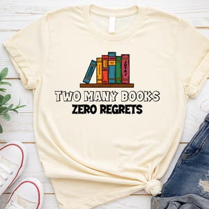 May include: Cream-colored t-shirt featuring a graphic of colorful books on a shelf. The text "TWO MANY BOOKS ZERO REGRETS" is printed below the books in black. The shirt is paired with blue denim shorts and white sneakers.