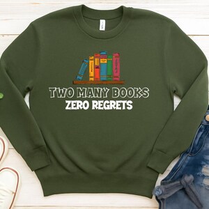 May include: An olive green sweatshirt with the text "TWO MANY BOOKS ZERO REGRETS" in white. The graphic features a shelf with a stack of colorful books. The sweatshirt is paired with blue jeans and white sneakers.
