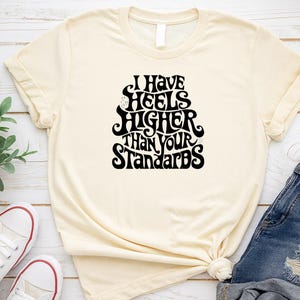 May include: Cream-colored t-shirt with the text "I Have Heels Higher Than Your Standards" in black. The shirt is paired with blue denim shorts and white sneakers with red accents.