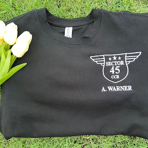 May include: Black crew neck sweatshirt with a white graphic on the chest that reads "SECTOR 45 CCR" with a wing design and the name "A. WARNER" below. The sweatshirt is lying on green grass next to a bouquet of white tulips.