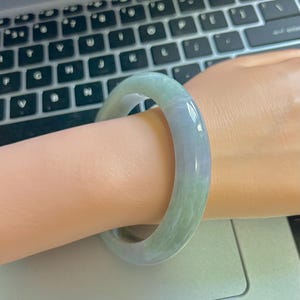 Op de afbeelding: Een jade armband in groen- en lavendeltinten wordt om een pols gedragen. Het gladde, gepolijste oppervlak van de armband reflecteert licht en benadrukt de natuurlijke kleurvariaties. De armband is een klassiek sieraad.