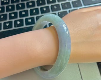 May include: A jade bangle bracelet in shades of green and lavender is worn on a wrist. The smooth, polished surface of the bracelet reflects light, highlighting its natural color variations. The bracelet is a classic piece of jewelry.