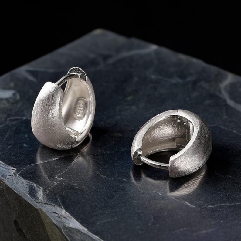 Pode incluir: Um par de brincos de argola em prata com acabamento mate e texturizado. Os brincos apresentam um design espesso e arredondado com fecho de dobradi&ccedil;a. O interior de uma argola mostra uma pequena marca que indica a qualidade do metal.