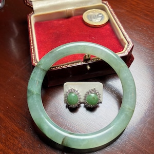 Op de afbeelding: Een lichtgroene jade armband en bijpassende oorknopjes. De armband is cirkelvormig. De oorbellen hebben een groene middensteen omringd door kleine, heldere stenen. De sieraden worden gepresenteerd op een houten oppervlak.
