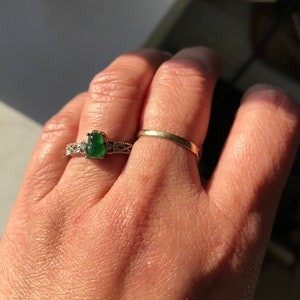 May include: A hand wearing two rings. The ring on the index finger is a thin gold band. The ring on the middle finger has a large green gemstone surrounded by a silver band with intricate details.