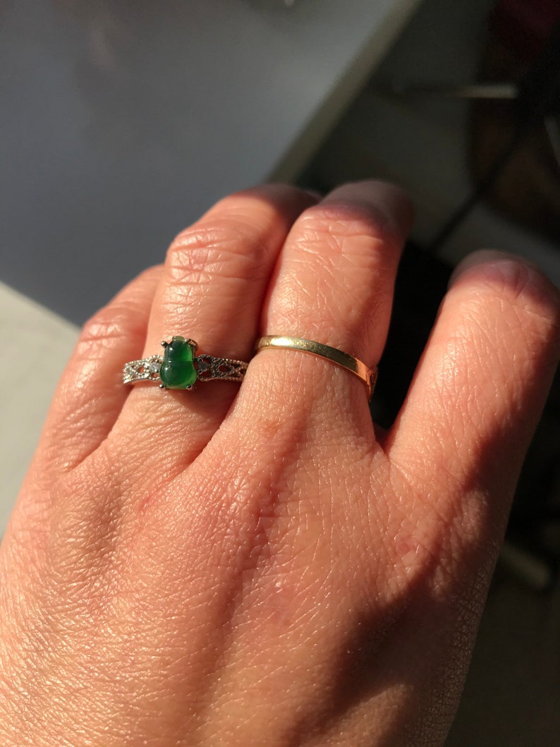 May include: A hand wearing two rings. The ring on the index finger is a thin gold band. The ring on the middle finger has a large green gemstone surrounded by a silver band with intricate details.