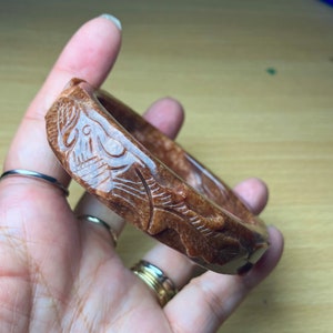 Op de afbeelding: Een bruine gegraveerde jade armband met ingewikkelde bloemmotieven. De armband is gemaakt van een doorschijnend materiaal met een roodachtige tint.