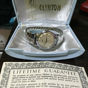 Può includere: Un orologio Clinton vintage nella sua scatola azzurra originale. L'orologio ha un cinturino argentato e un quadrante color crema con numeri e lancette nere. La scatola è aperta, rivelando l'orologio e un documento di garanzia.