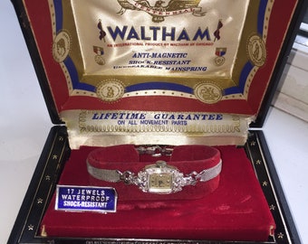 Puede incluir: Reloj Waltham vintage en una caja de presentación negra. El reloj tiene una correa de malla plateada y una esfera cuadrada con un bisel con forma de diamante. El interior de la caja es de terciopelo rojo con detalles dorados y azules. La caja dice "Waltham" y "17 Jewels Waterproof Shock-Resistant."
