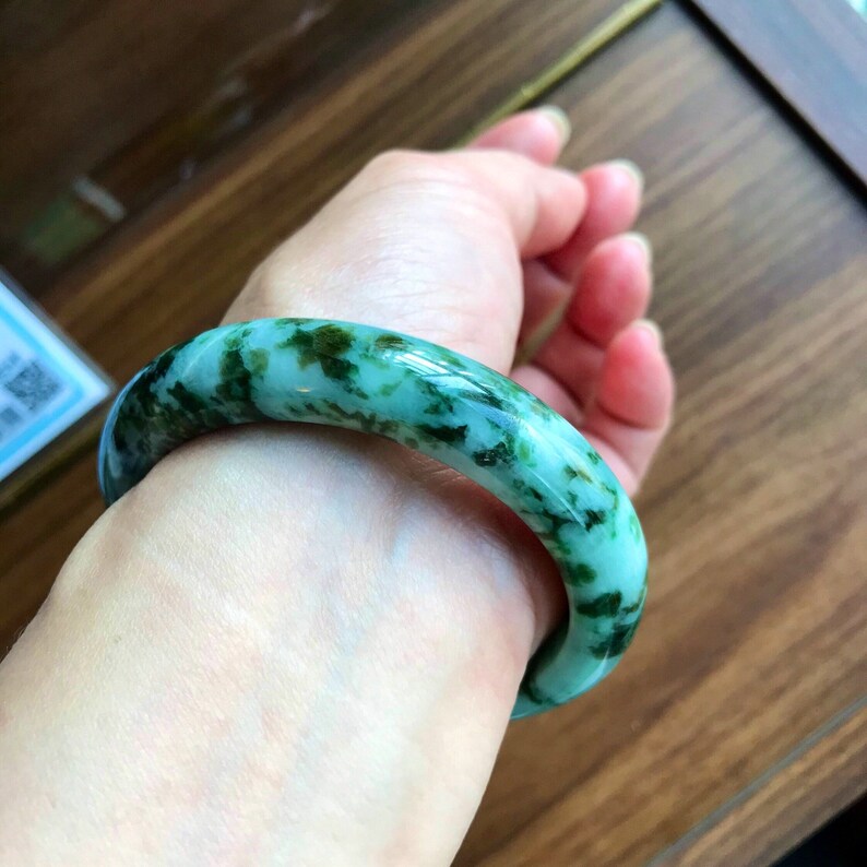 May include: A green jade bangle bracelet with a mottled pattern. The bracelet is worn on a person's wrist.