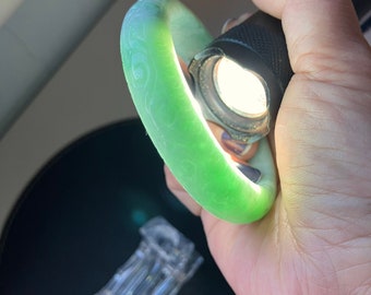 May include: A green jade bangle with intricate carvings. The bangle is being held in a hand and illuminated by a flashlight.