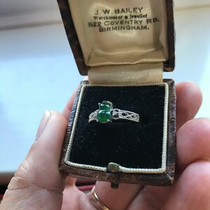 May include: A green jade ring with a silver band and intricate detailing, in a brown velvet ring box. The box is labeled "J.W. BAILEY Watchmaker & Jeweller 522 COVENTRY RD. BIRMINGHAM."