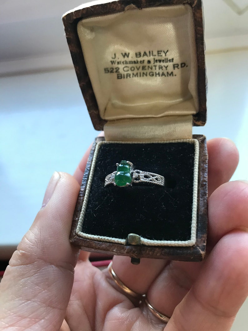 May include: A green jade ring with a silver band and intricate detailing, in a brown velvet ring box. The box is labeled "J.W. BAILEY Watchmaker & Jeweller 522 COVENTRY RD. BIRMINGHAM."
