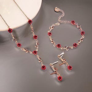May include: A gold-coloured jewellery set comprising a necklace, bracelet, and earrings. Each item is decorated with red rose-shaped details and small, sparkling embellishments. The set is presented on a grey surface.