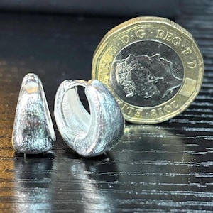 Pode incluir: Dois brincos de argola prateados com um acabamento texturizado e mate. Um brinco est&aacute; posicionado na vertical, enquanto o outro est&aacute; de lado. Uma moeda brit&acirc;nica de uma libra est&aacute; colocada ao lado para mostrar a escala do objeto.
