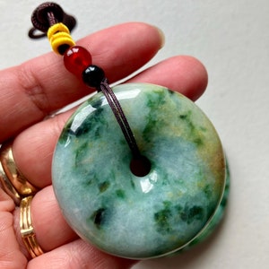 May include: A green jade pendant in the shape of a donut with a brown cord attached. The pendant has a light green color with darker green swirls and speckles.
