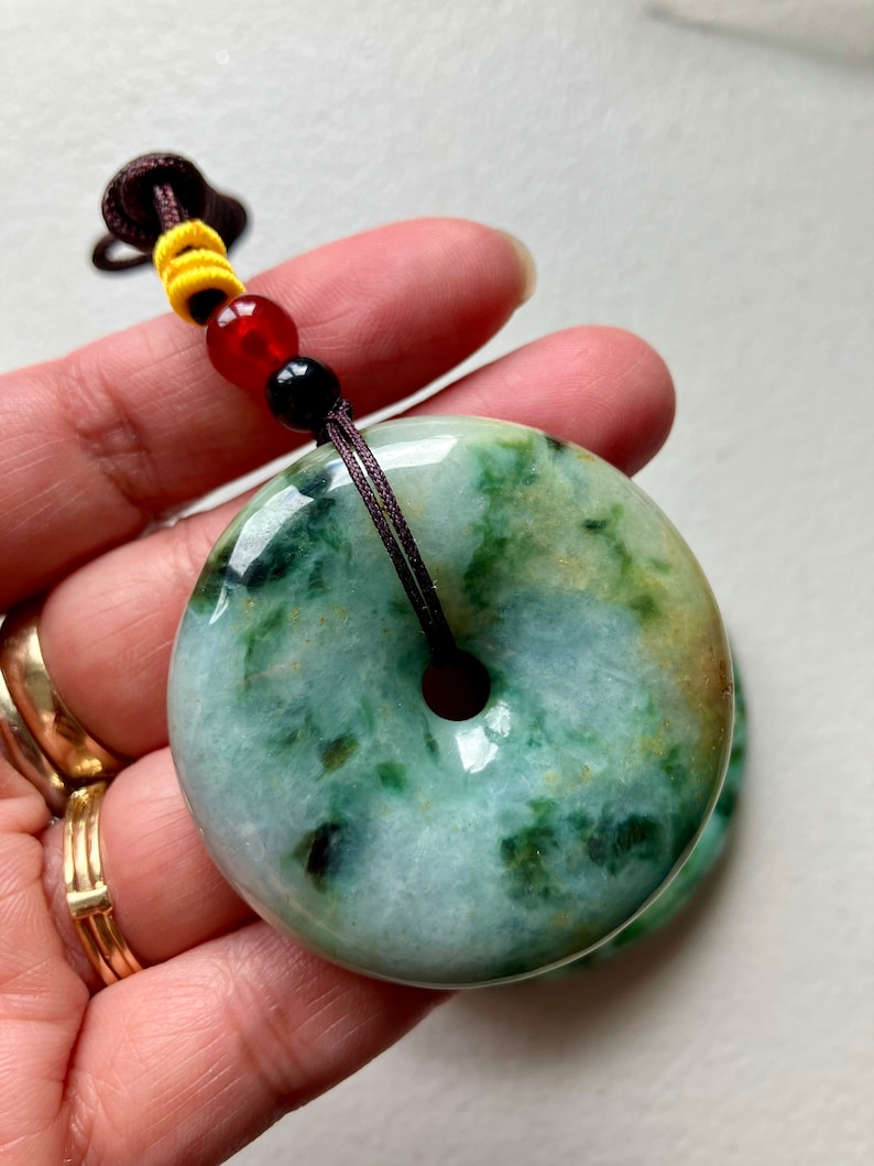 May include: A green jade pendant in the shape of a donut with a brown cord attached. The pendant has a light green color with darker green swirls and speckles.