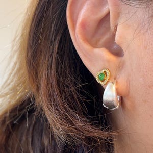 Pode incluir: Um brinco de pino dourado em forma de cora&ccedil;&atilde;o com uma pequena pedra verde circular rodeada por min&uacute;sculos cristais transparentes, combinado com um brinco de argola grosso em tom de prata escovada.