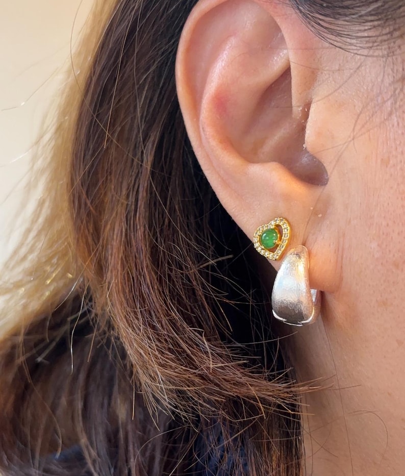Pode incluir: Um brinco de pino dourado em forma de cora&ccedil;&atilde;o com uma pequena pedra verde circular rodeada por min&uacute;sculos cristais transparentes, combinado com um brinco de argola grosso em tom de prata escovada.