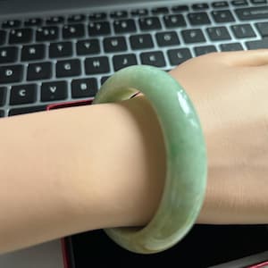 May include: A jade bangle bracelet in shades of green and yellow is worn on a wrist. The smooth, polished surface of the bracelet reflects light. The background includes a laptop keyboard and a red box.