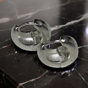 Pode incluir: Um par de brincos de argola em prata com um design espesso e arredondado com um acabamento texturizado e mate. Os brincos s&atilde;o constru&iacute;dos com um mecanismo de pino articulado que prende a argola, exibindo um brilho met&aacute;lico e um perfil s&oacute;lido.