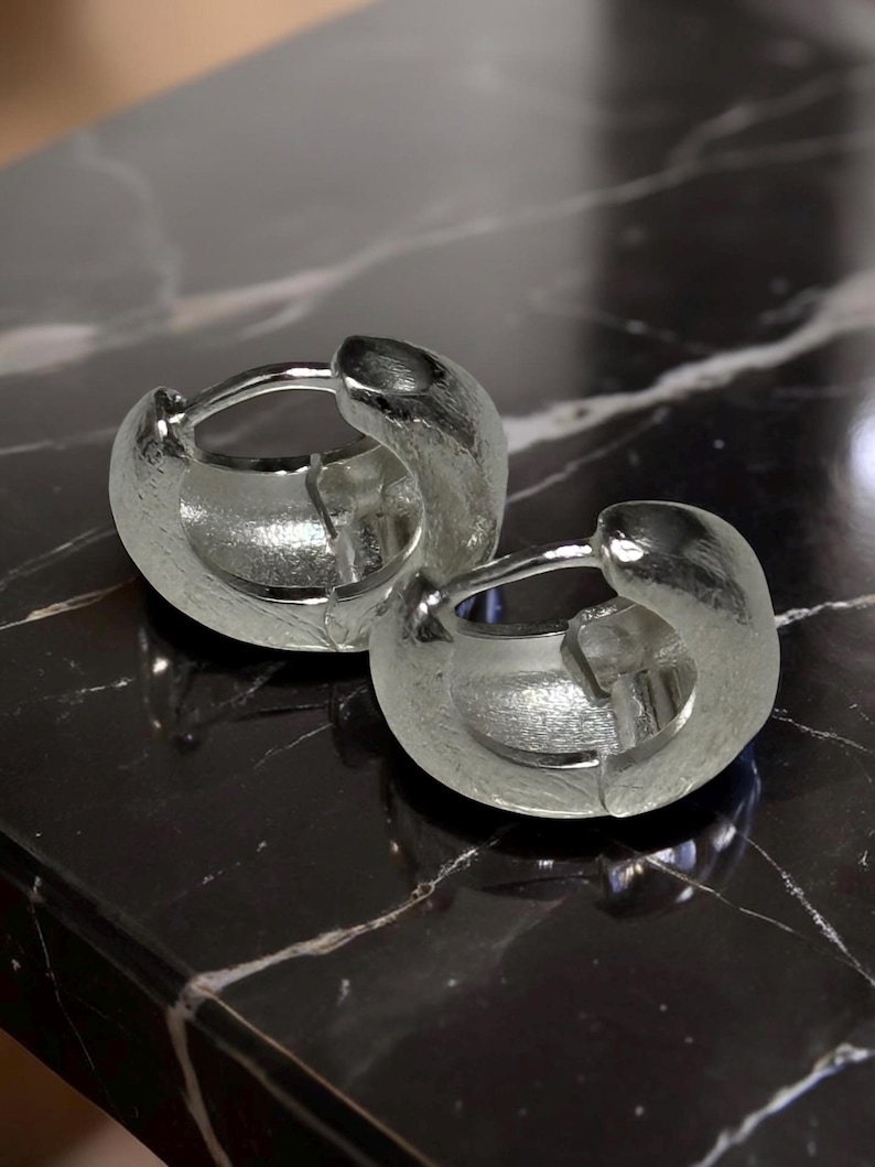 Pode incluir: Um par de brincos de argola em prata com um design espesso e arredondado com um acabamento texturizado e mate. Os brincos s&atilde;o constru&iacute;dos com um mecanismo de pino articulado que prende a argola, exibindo um brilho met&aacute;lico e um perfil s&oacute;lido.