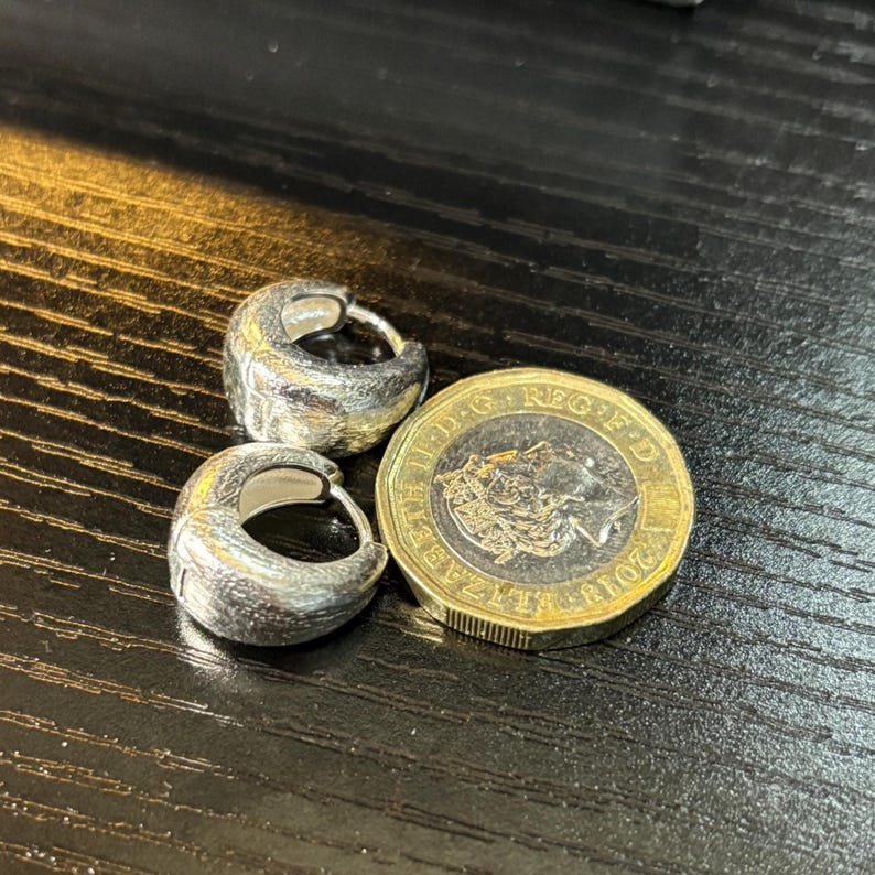 Pode incluir: Dois pequenos brincos de argola em prata com um acabamento texturizado e mate. Os brincos est&atilde;o posicionados ao lado de uma moeda brit&acirc;nica de uma libra, que serve como refer&ecirc;ncia de escala para o seu tamanho.