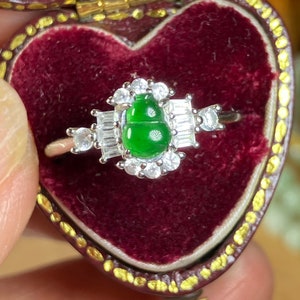 Può includere: Un anello d'argento con una vivace pietra verde a forma di zucca, circondata da scintillanti pietre bianche. L'anello è presentato su uno sfondo di velluto rosso scuro, evidenziando l'eleganza e il design del gioiello.