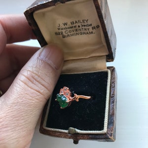 May include: A green gemstone ring with a floral design set in a rose gold band. The ring is in a small, brown, wooden box with a white interior. The box has the text "J.W. BAILEY Watchmaker & Jeweller 522 COVENTRY RD BIRMINGHAM." printed on the inside.