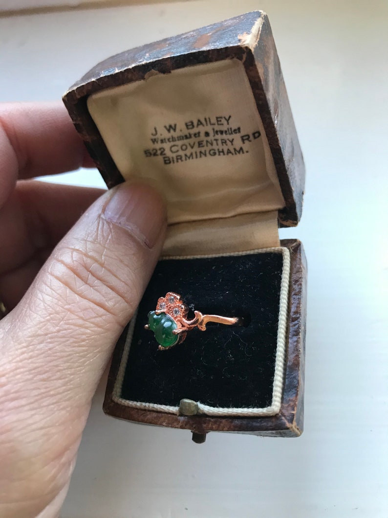 May include: A green gemstone ring with a floral design set in a rose gold band. The ring is in a small, brown, wooden box with a white interior. The box has the text "J.W. BAILEY Watchmaker & Jeweller 522 COVENTRY RD BIRMINGHAM." printed on the inside.