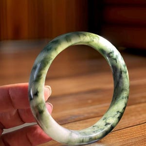 Puede incluir: Un brazalete de jade verde con vetas blancas y negras. El brazalete es liso y pulido.