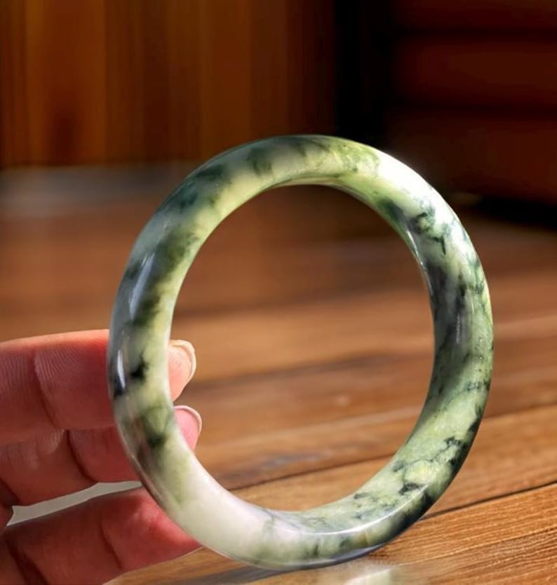 Puede incluir: Un brazalete de jade verde con vetas blancas y negras. El brazalete es liso y pulido.