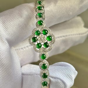 Op de afbeelding: Zilveren armband met een bloemmotief, bezet met ronde, levendige groene edelstenen en kleine, heldere accentstenen. De armband heeft een scharniersluiting en is geplaatst tegen een witte achtergrond.