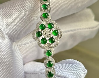 May include: A silver bracelet featuring a floral design with round, vibrant green gemstones and small, clear accent stones. The bracelet has a hinged clasp and is set against a white background.