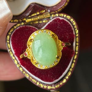 May include: A gold ring featuring a large, oval, light green cabochon stone at the center, surrounded by a halo of small clear stones. The setting includes intricate gold filigree details and two small, round, red accent stones on the sides.