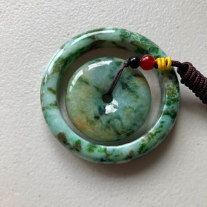 May include: A jade pendant with a green and brown marbled pattern. The pendant is a round disc with a hole in the center. It is attached to a jade bangle with a similar marbled pattern.