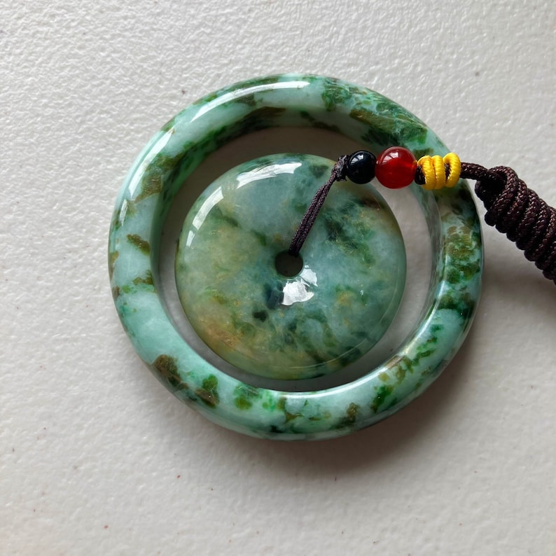 May include: A jade pendant with a green and brown marbled pattern. The pendant is a round disc with a hole in the center. It is attached to a jade bangle with a similar marbled pattern.
