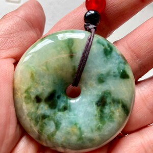 May include: A green jade pendant with a hole in the center, hanging from a brown cord with black and red beads. The pendant is a round, flat disc with a mottled green and white pattern.