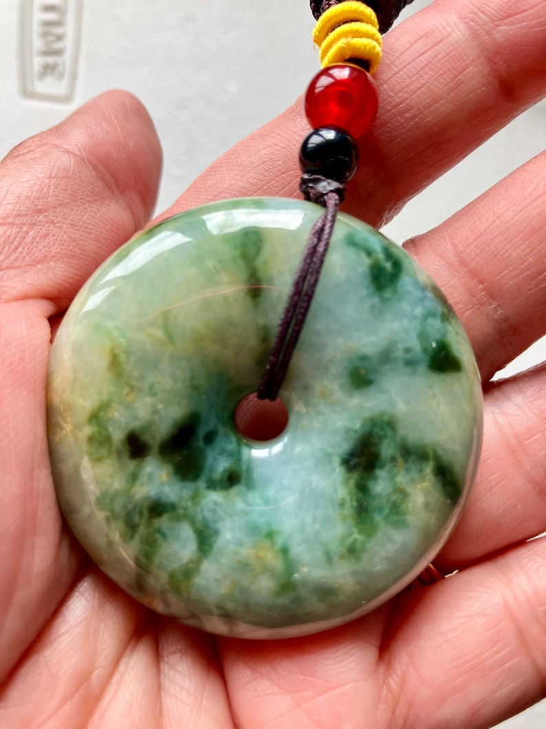 May include: A green jade pendant with a hole in the center, hanging from a brown cord with black and red beads. The pendant is a round, flat disc with a mottled green and white pattern.
