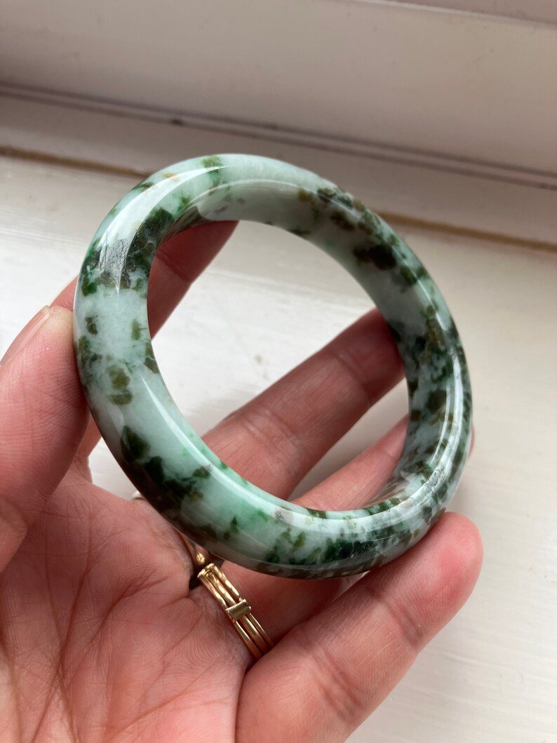 May include: A green jade bangle bracelet with a mottled pattern. The bracelet is smooth and polished.