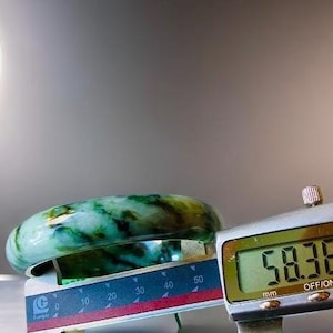 Puede incluir: Un brazalete de jade con vetas verdes y blancas, medido con un calibre digital que muestra 58,36 mm.