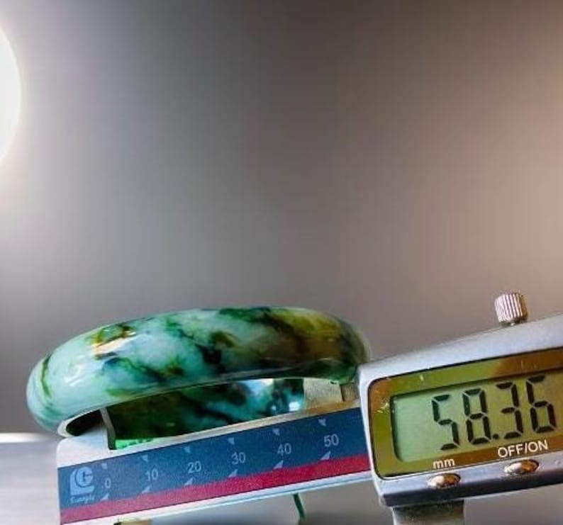 Puede incluir: Un brazalete de jade con vetas verdes y blancas, medido con un calibre digital que muestra 58,36 mm.