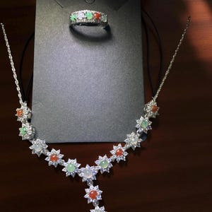 Könnte beinhalten: Ein silbernes Halsketten- und Ring-Set. Die Halskette besteht aus sternförmigen Anhängern mit roten, grünen und weißen Edelsteinen, akzentuiert durch klare Steine. Der Ring hat ein ähnliches Design mit den gleichen farbigen Edelsteinen.