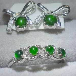 May include: A set of jewellery consisting of two bow-shaped stud earrings and a ring. Each piece is crafted from silver-toned metal, accented with small clear stones, and features round, smooth, deep green cabochon-style stones as the focal point.