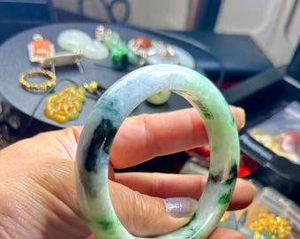 May include: A jade bangle bracelet with a mix of green, white, and black colors. The bracelet is held up in front of other jewelry pieces. The jade is polished and smooth, with a marbled appearance. The bangle is a circular shape.