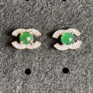 May include: A pair of stud earrings featuring a central round green cabochon stone set within a silver-toned metal frame shaped like two interlocking C letters, with small clear pavé-set stones embedded along the curved metal surfaces.