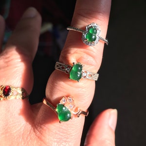 May include: Three jade rings with silver and gold settings. The jade stones are green and shaped like gourds. The rings are on a finger.